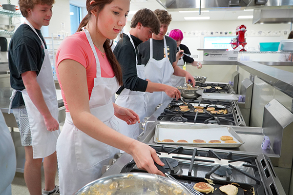 several students in aprons flip pancakes on griddles